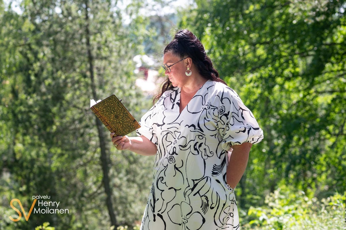 Jarna Pihlajamäki lukee kirjaa luonnon keskellä valkoisessa mekossaan.