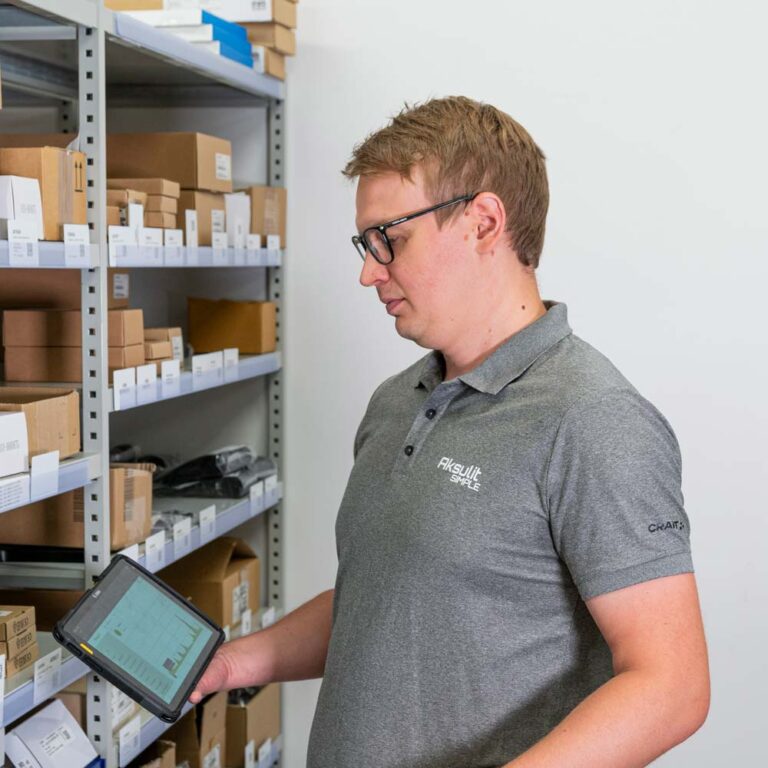 Mies selaa varastossa tablettia, jossa näkyy tuotehallinnan näkymä hyllyjen ja pakkausten edessä.