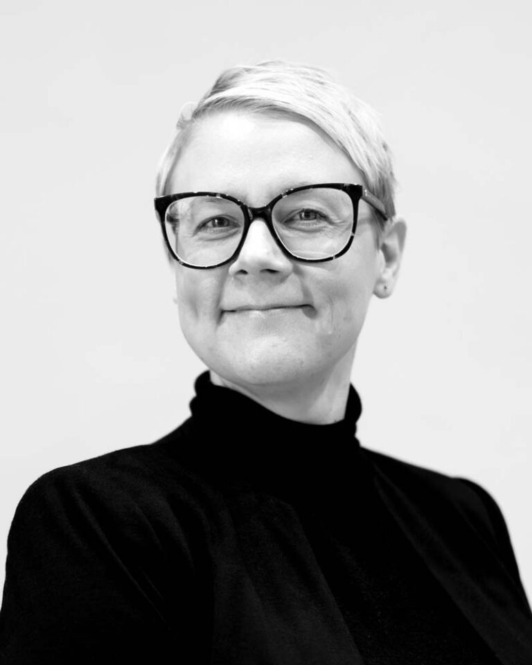 Mustavalkoinen kasvokuva lyhythiuksisesta henkilöstä, jolla on paksut mustat silmälasit ja musta poolopaita.